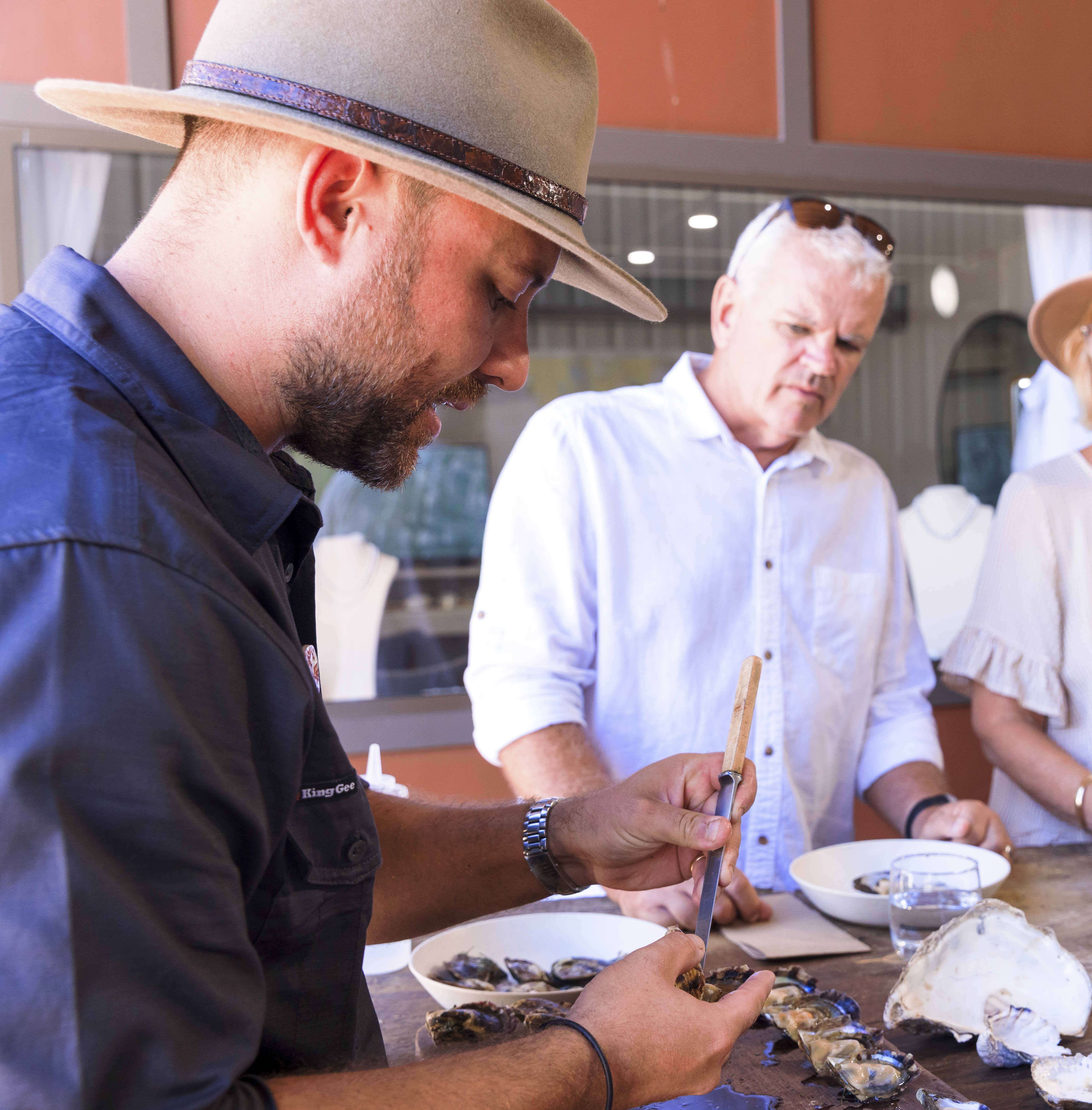 A man shucking an oyster, with a man in the background watching.